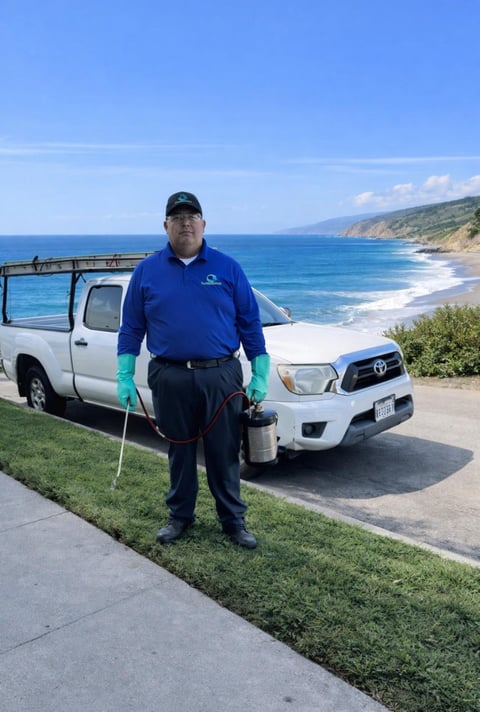 Pest control technician in blue uniform holding equipment standing in front of service vehicles overlooking a coastal beach with cliffs