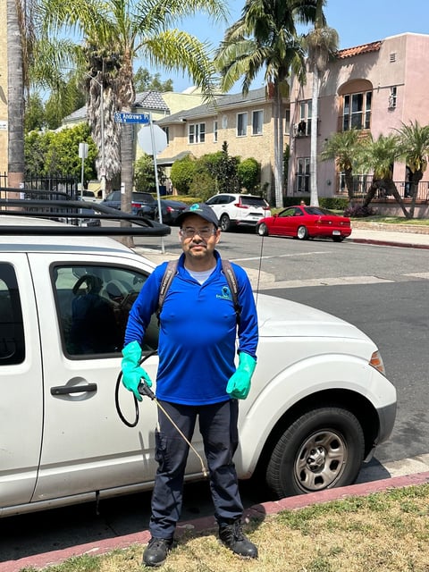 Worker in blue shirt and green gloves standing with pest control equipment next to white vehicle