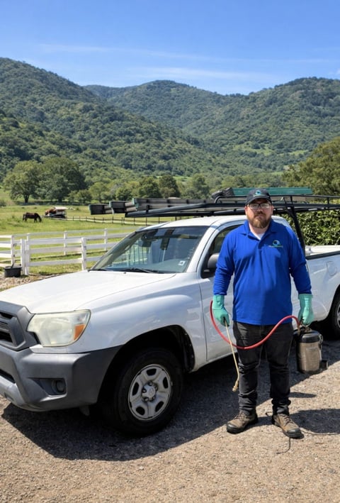 Man in blue work shirt standing beside white pickup truck on rural property with green mountains and farm fencing in background